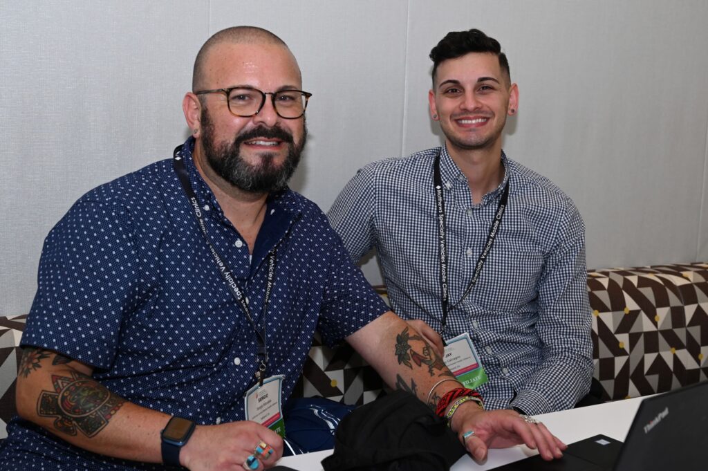 A man with a beard wearing glasses and a short sleeved dark blue shirt and a man with blue checkered long sleeved shirt sit on a couch and smile at the camera