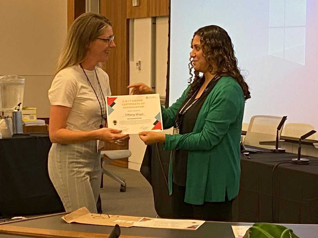 A woman with long blonde hair wearing glasses and light clothing is handed a certificate by a woman with long wavy dark hair wearing a green sweater over a black shirt.